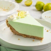 A slice of creamy lime Jello pie with a graham cracker crust, topped with whipped cream and a lime wedge, served on a white plate.